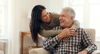Happy senior Caucasian man and young Asian woman embracing and laughing together on couch in bright living room showing intergenerational love and family bonding.