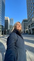 Mature woman with long gray hair wearing dark winter jacket standing in urban plaza surrounded by modern skyscrapers on clear sunny day, looking upward with positive expression.