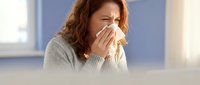 Woman with reddish-brown curly hair, appearing to be in her 30s-40s of mixed ethnicity, blowing her nose into a tissue while sitting indoors. She wears a light gray knit sweater and has a facial expression suggesting illness or allergies. Bright window and blue wall visible in background. Suitable for healthcare, wellness, and medical content.