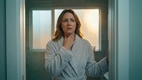 Middle-aged Caucasian woman in bathrobe touching throat with concerned expression standing in bathroom doorway during morning light for health concepts.