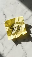 Crumpled yellow sticky note with handwritten word Sorry on white marble surface in natural sunlight, conveying apology and regret.