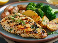 Grilled herb-seasoned chicken breast with rosemary garnish, served alongside roasted potatoes and steamed broccoli on ceramic plate. Fresh citrus and herbs enhance presentation.