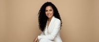 Professional Hispanic businesswoman in white blazer suit smiling confidently against neutral beige background for corporate marketing and business presentations.