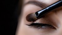 Close up of eye makeup application showing professional brush applying shimmering bronze eyeshadow on model's eyelid against dark background.