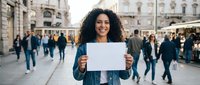 Young mixed-race woman with curly hair holding blank white paper sign in busy urban plaza with historic architecture and crowd of people walking.