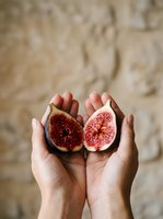 Two halves of a fresh fig held in open palms against a neutral textured background, showcasing the vibrant red interior and seeds for food, nutrition, and healthy eating concepts.