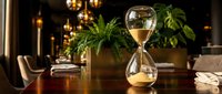 Elegant hourglass with golden sand on wooden table in luxurious office interior with warm lighting and green plants creating sophisticated atmosphere.