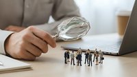Professional businessman examining miniature business team figures with magnifying glass for recruitment and talent acquisition analysis.
