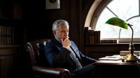 Senior businessman in navy suit sitting in leather chair at wooden desk in traditional office library, contemplating with hand to chin, natural light from arched window, professional workspace for corporate leadership and executive portraits.