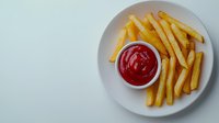 Crispy golden french fries served on white ceramic plate with bright red ketchup dipping sauce, shot from above on light background. For restaurant menu or food blog.