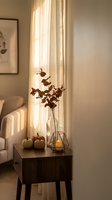 Cozy autumn home interior with dried branches in vase, pumpkins and candle on wooden table by window with warm sunlight streaming through curtains.