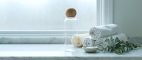 Spa wellness setup with rolled towels, essential oil bottle and eucalyptus on white bathroom counter near window for relaxation and self-care rituals.