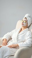 Woman relaxing in white bathrobe with cucumber eye mask and towel on head during spa treatment at home for skincare and wellness routine.