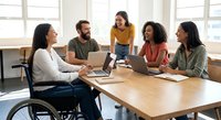Diverse business team meeting with disabled woman in wheelchair collaborating on project in modern office workspace with laptops and documents.