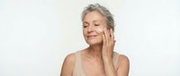Mature Caucasian woman with gray hair applying skincare cream to face with gentle touch, eyes closed in peaceful expression for anti-aging beauty routine.