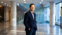Confident middle-aged Caucasian businessman in navy suit standing in modern corporate lobby with marble columns and large windows during daytime.