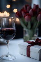 Glass of red wine with romantic evening setting, gift box with burgundy ribbon and red tulips in background against soft bokeh lights. Elegant Valentine mood.