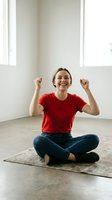 Happy young woman celebrating success sitting cross-legged on yoga mat in bright modern room with raised fists expressing joy and achievement.