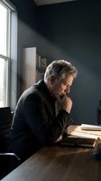 Mature man with gray hair and Caucasian ethnicity sitting at wooden desk in dark office, hand to chin in thoughtful pose, natural window light, contemplative mood.