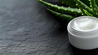 Natural aloe vera moisturizing cream in white jar on dark slate background. Organic skincare product with fresh green aloe plant leaves for beauty routine.