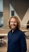 Smiling middle-aged Caucasian businessman with curly blonde hair and beard in navy shirt standing confidently in modern office workspace.