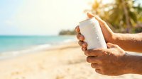 Person applying sunscreen lotion on arm at tropical beach with turquoise water and palm trees, sun protection skincare product for summer vacation and outdoor activities.