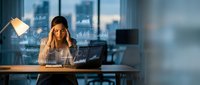 Stressed businesswoman working late at office desk with laptop and financial data charts displayed on screen in modern corporate environment.