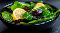 Fresh green spinach salad with sliced lemon and dark purple beets served in black ceramic bowl on dark background, close up view of healthy vegetarian meal.