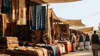 Traditional Middle Eastern carpet market with handwoven rugs and textiles displayed under canvas awnings, vendors in cultural dress browsing colorful merchandise.