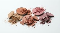 Broken makeup powder compacts in warm nude and pink tones scattered with crumbled cosmetic fragments on white background for beauty concepts.