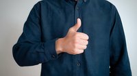 Man in navy blue button-up shirt showing thumbs up gesture against neutral background, conveying approval, satisfaction, or positive feedback.