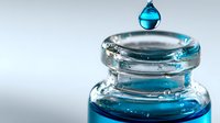 Blue water droplet suspended above glass container with turquoise liquid, capturing moment before impact in high-speed macro photography.