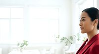 Mature Asian woman in red blazer sitting by bright window with potted plants, looking thoughtful in modern office or home workspace.