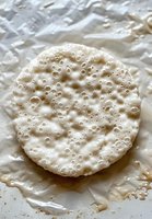 Round rice cake with airy bubbled texture on white paper, macro food photography showing detailed porous surface and natural grain pattern.