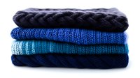 Neatly folded stack of cozy winter sweaters in shades of navy blue and turquoise, showcasing different knit patterns and textures against white background.