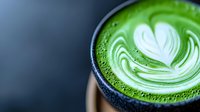 Vibrant green matcha latte with white heart-shaped latte art foam design in dark ceramic cup, close-up view showing rich texture and contrast.