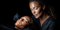 Professional African American female tattoo artist working on client's eyebrow in modern studio setting with focused lighting and artistic atmosphere.