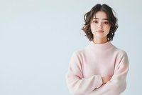 Young Asian woman with short wavy hair wearing pink sweater standing with crossed arms against white background, expressing confidence and friendly attitude.
