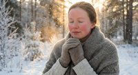 Young Caucasian woman in knit sweater and gloves standing in snowy winter forest, eyes closed, warming hands in golden sunlight.