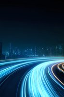 Futuristic night cityscape with glowing blue light trails from traffic creating dynamic curved lines against dark urban skyline with illuminated skyscrapers.