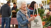 Senior woman with curly gray hair shopping at farmers market with eco-friendly tote bag, smiling while selecting fresh organic vegetables and produce.