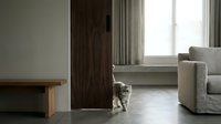 Small tabby kitten walking across modern minimalist living room with wooden bench, beige sofa and natural light from window creating cozy domestic scene.