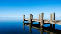 Weathered wooden pier extending into calm blue water under clear sky, suitable for travel, nature, and tranquility concepts.