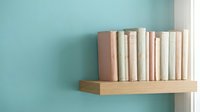 Pastel colored books arranged on wooden floating shelf against turquoise wall creating minimalist home decor and organized storage solution.