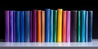 Row of colorful hardcover books standing upright on white shelf against black background, creating rainbow spectrum display of spines in varying heights.