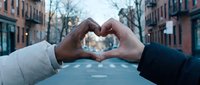 Two hands forming heart shape on urban street with brick buildings creating romantic gesture for love and connection concepts in city setting.