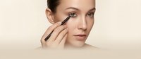 Young Caucasian woman applying eyeliner pencil near eye, close-up beauty makeup tutorial on beige background.