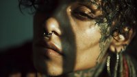 Close-up portrait of young woman with curly hair, nose ring, and earrings in dramatic lighting with shadow patterns on face. Artistic fashion photography.
