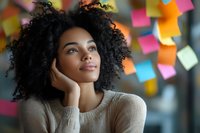 Young African American businesswoman with curly hair thinking creatively in modern office with colorful sticky notes on wall behind her.
