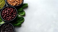 Variety of colorful legumes and beans in black bowls with fresh spinach leaves on light gray concrete background, border composition with copy space.